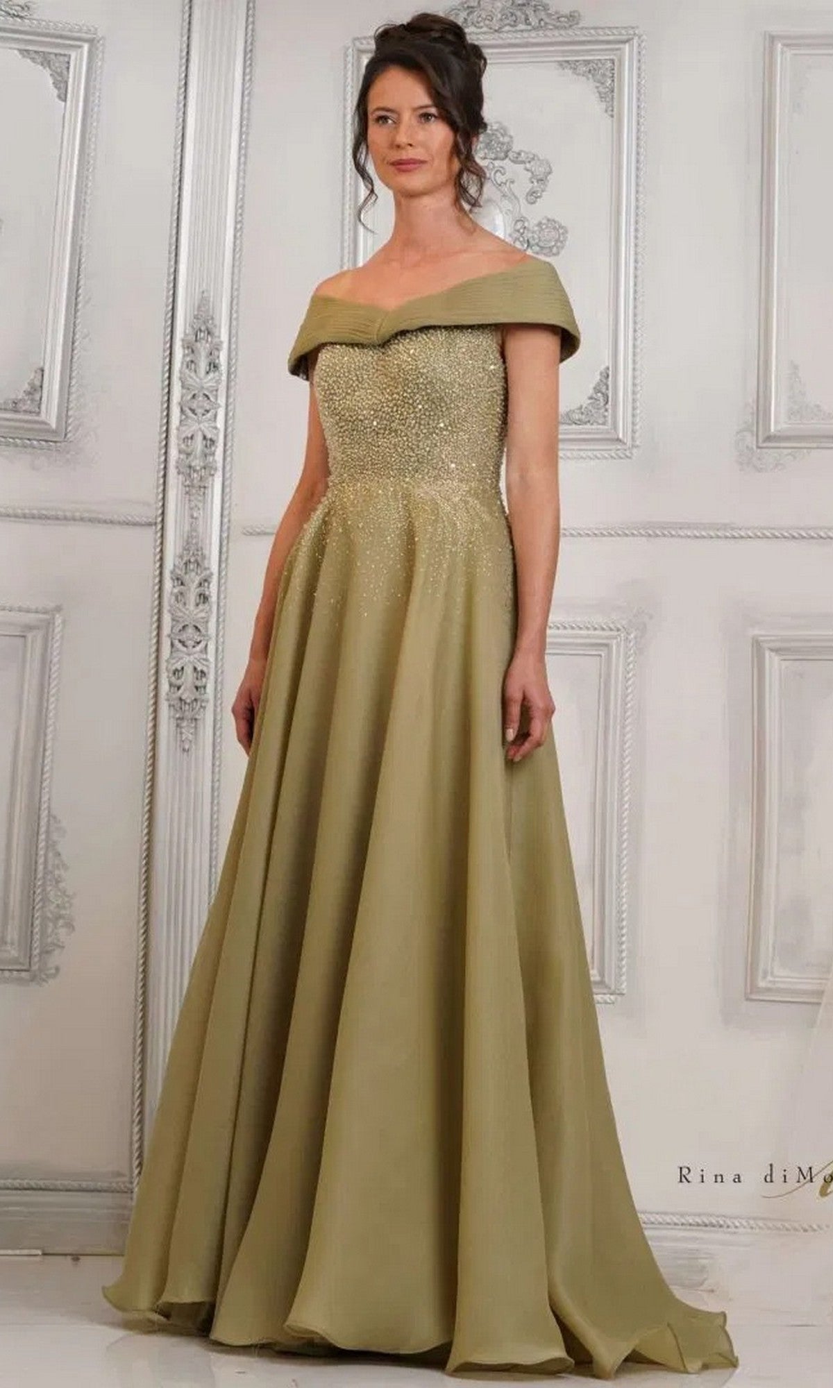 A woman stands gracefully against a decorative white background wearing the Rina Di Montella RD3102 Long Formal Dress in Olive Mist, featuring an off-the-shoulder beaded bodice and a flowing, floor-length A-line skirt.